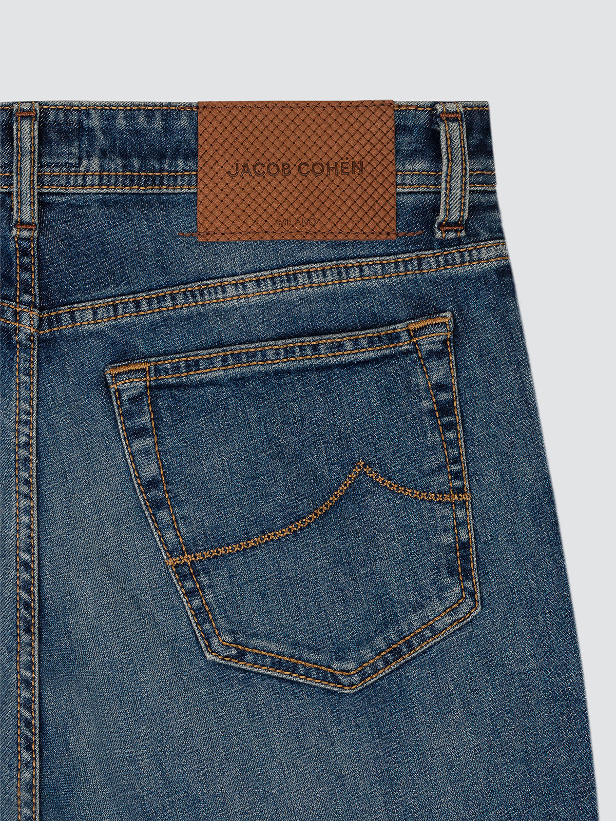 Close-up of the back pocket of medium blue Bard jeans, showing the rust brown leather patch with hot-stamped Jacob Cohën logo and the contrast arcuate stitching.