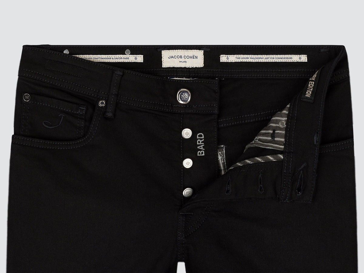 Close-up of black denim jeans showing the waistband, button fly, front pocket, and interior side pocket with a striped lining. Visible branding includes a central label reading JACOB COHEN and BARD on the fly.