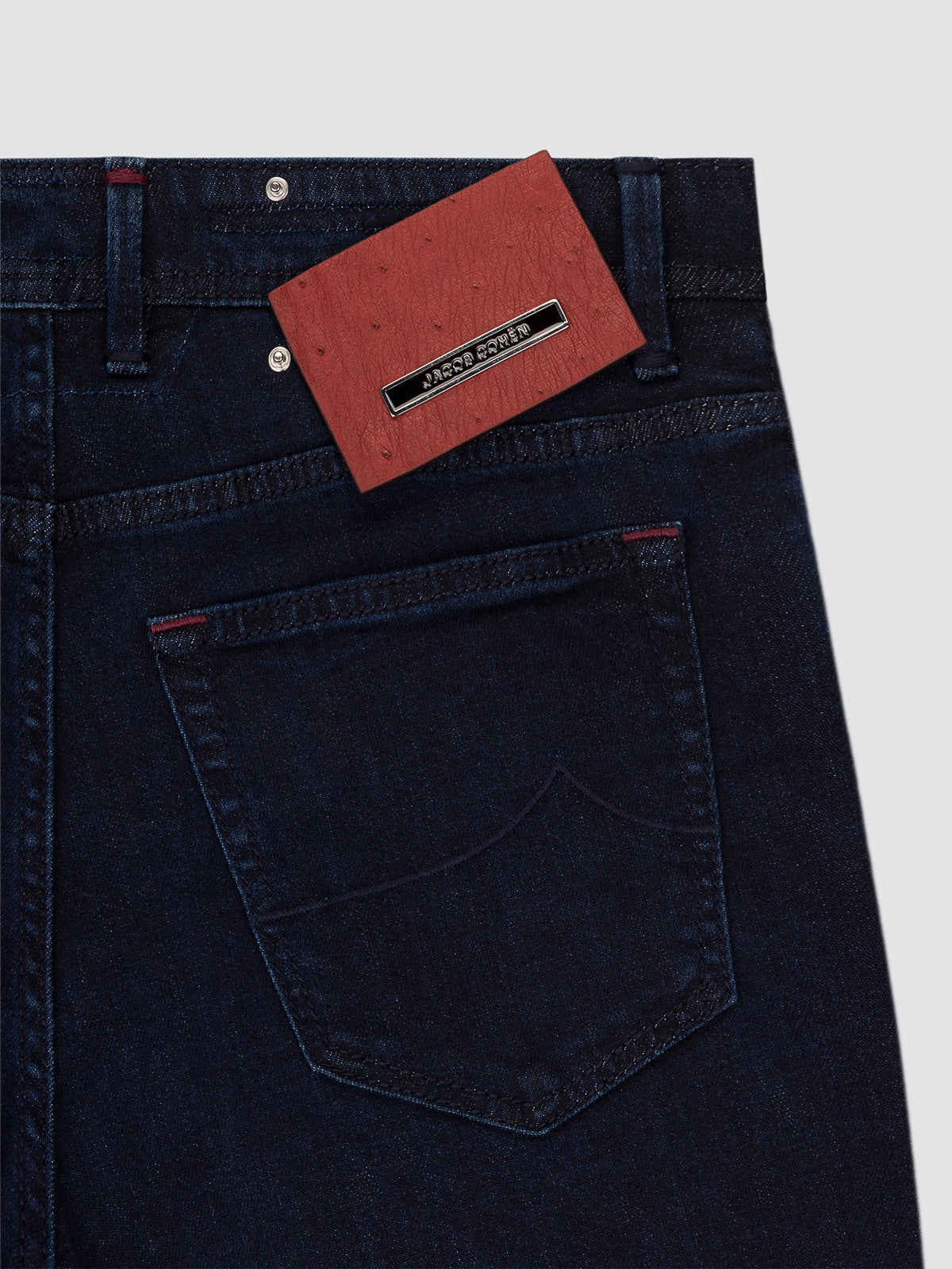 Close-up of the back waistband and pocket area of dark indigo denim jeans with a red paper brand tag attached near the belt loops on a light gray background.