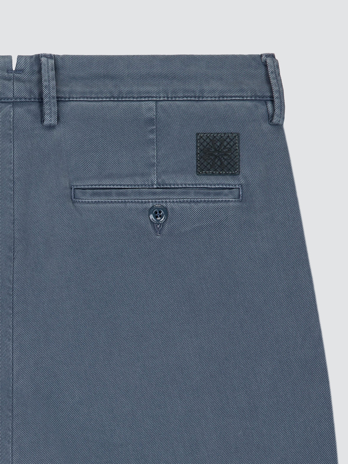 Close-up of the back waistband and rear pocket area of slate blue denim pants, showing belt loops, a buttoned welt pocket, and a small woven brand patch.