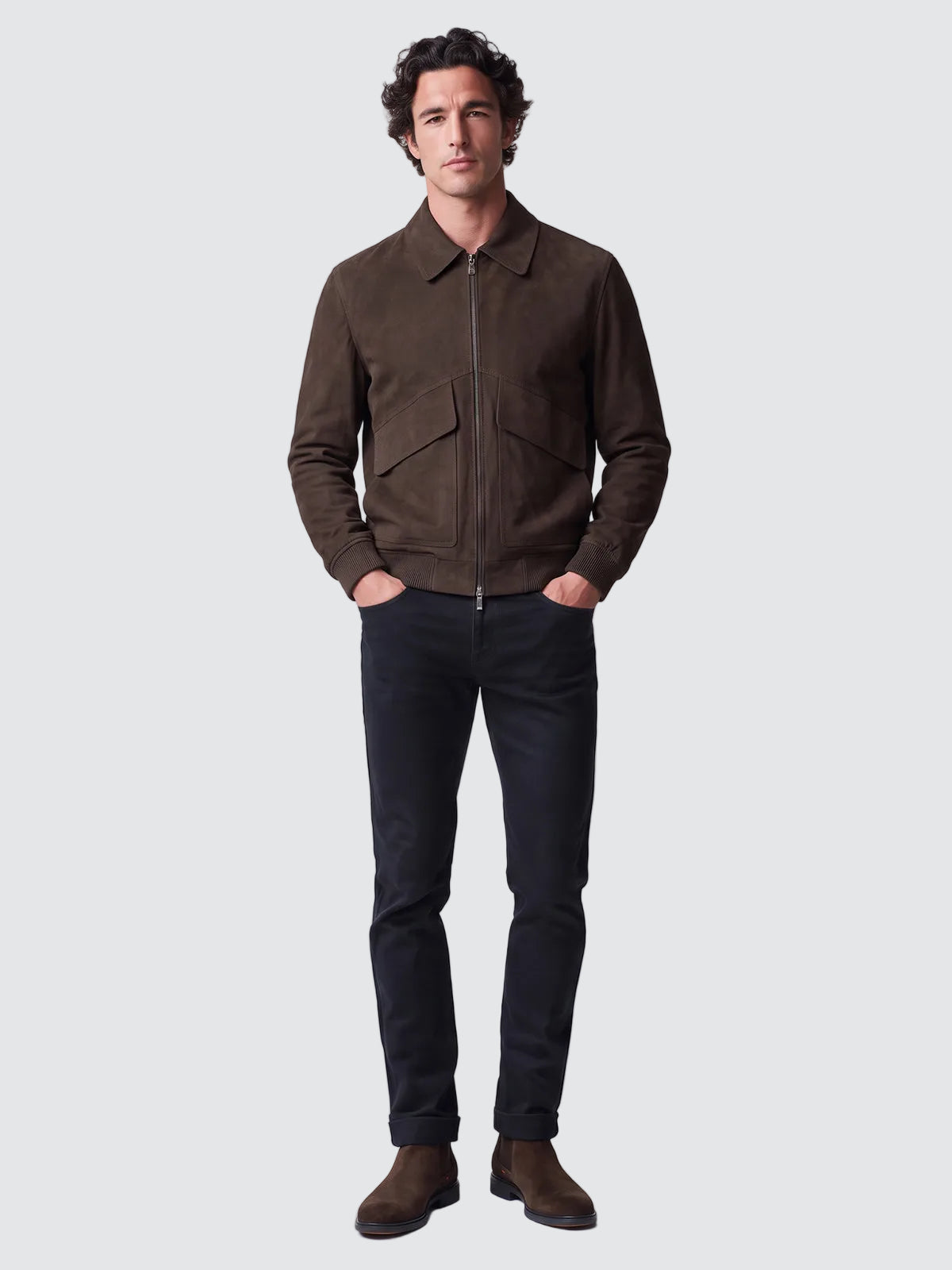 A man wearing a dark brown suede zip-up jacket, dark charcoal gray jeans, and brown suede boots, standing against a light gray studio background.