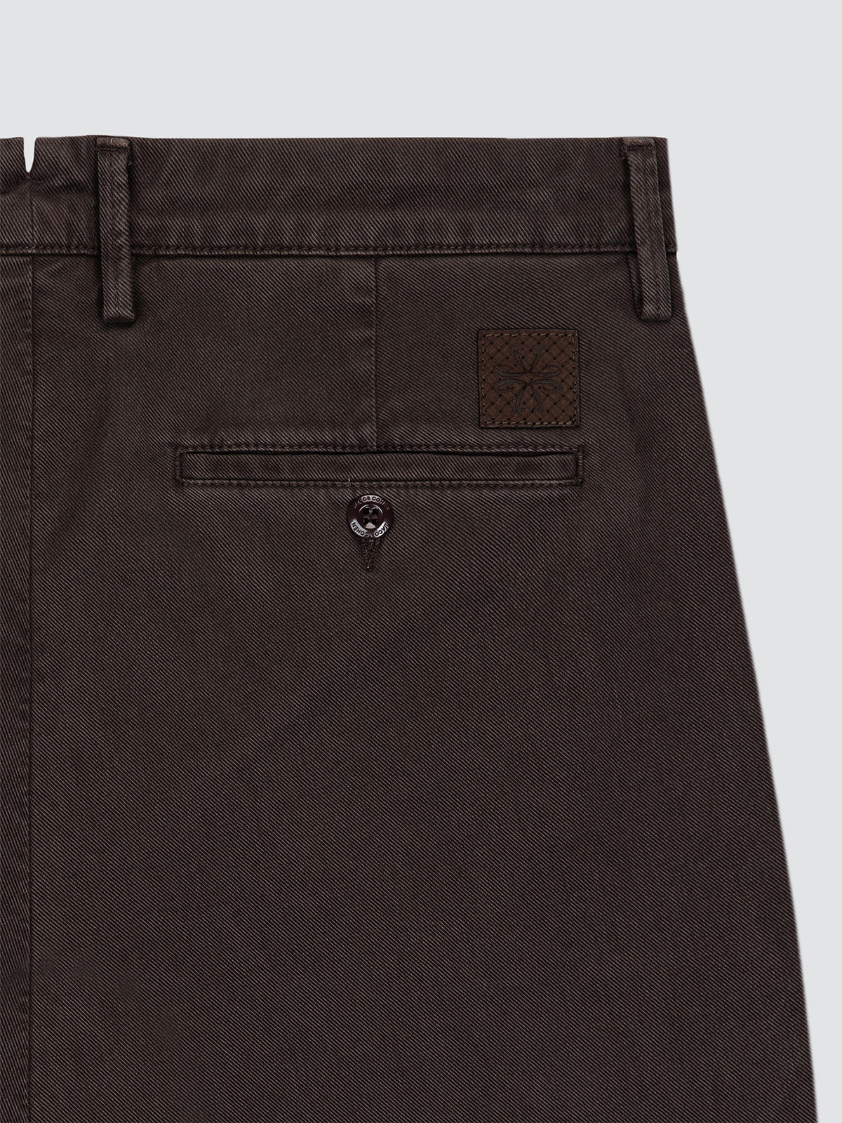 Close-up of the back waistband and rear pocket of dark brown trousers, showing belt loops, a buttoned welt pocket, and a small woven brand patch Jacob Cohën
