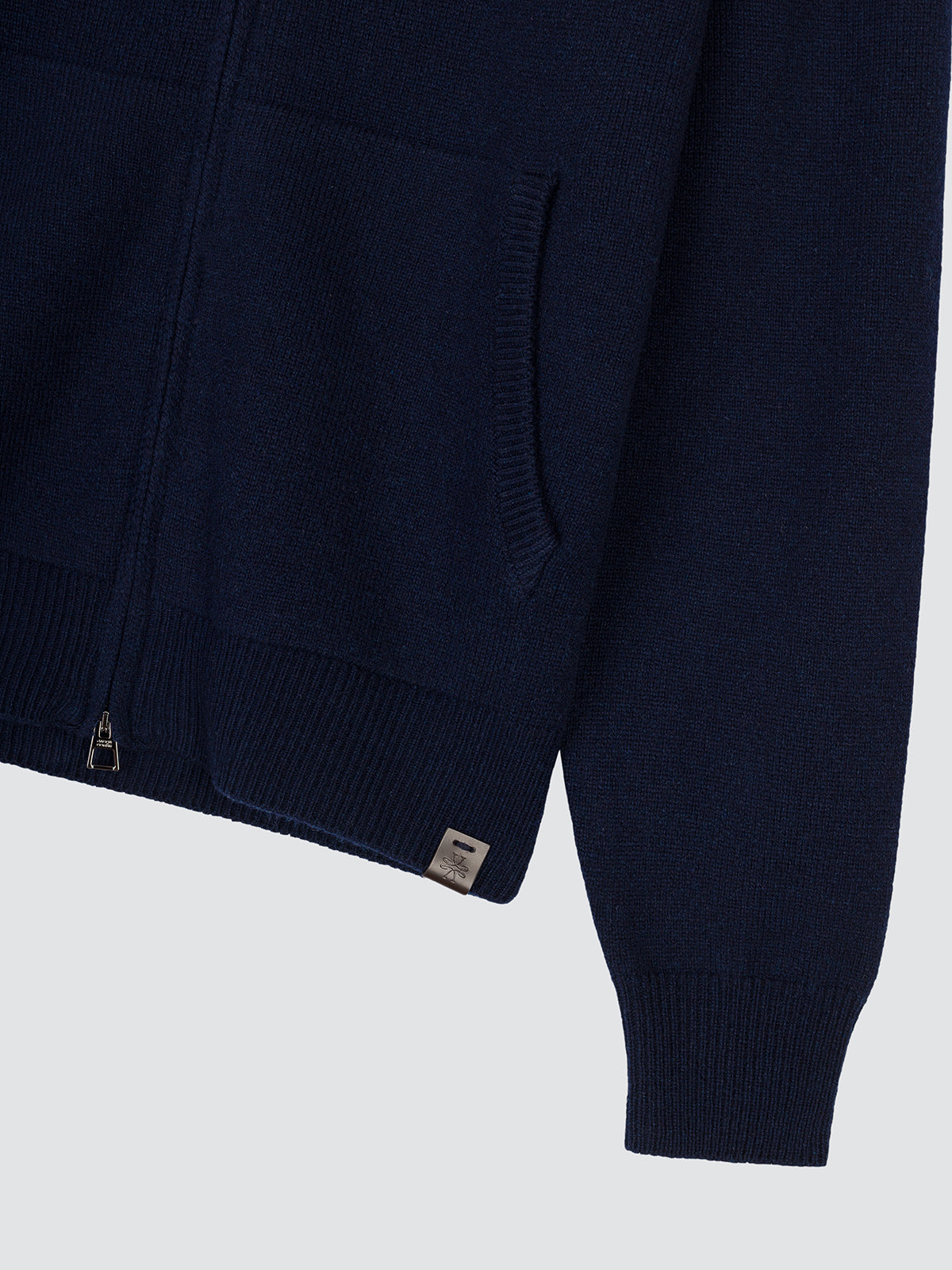 Close-up of a navy blue knit zip-up jacket, showing the sleeve, ribbed cuff, pocket, and a small square brand tag near the hem.