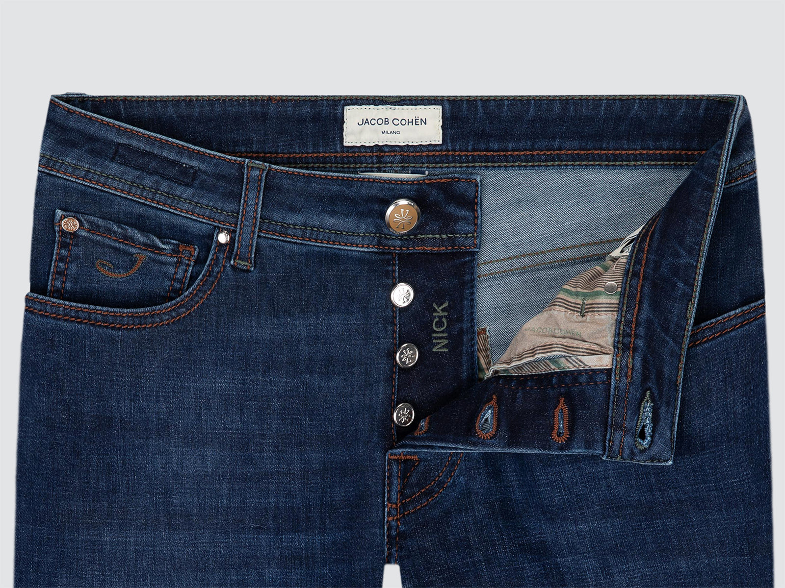 Close-up of dark blue denim jeans showing the waistband, front button closure, pocket, and inner lining details. A white waistband label reads JACOB COHEN model NICK