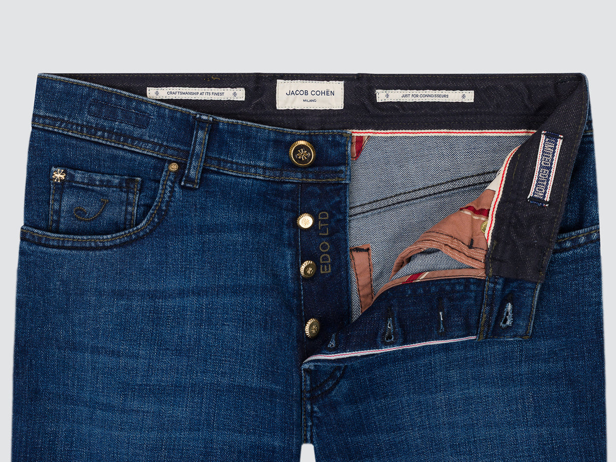 Close-up of a pair of blue denim jeans showing the waistband, front button closure with red line, pocket detail, and interior lining. The inner label reads Jacob Cohen.