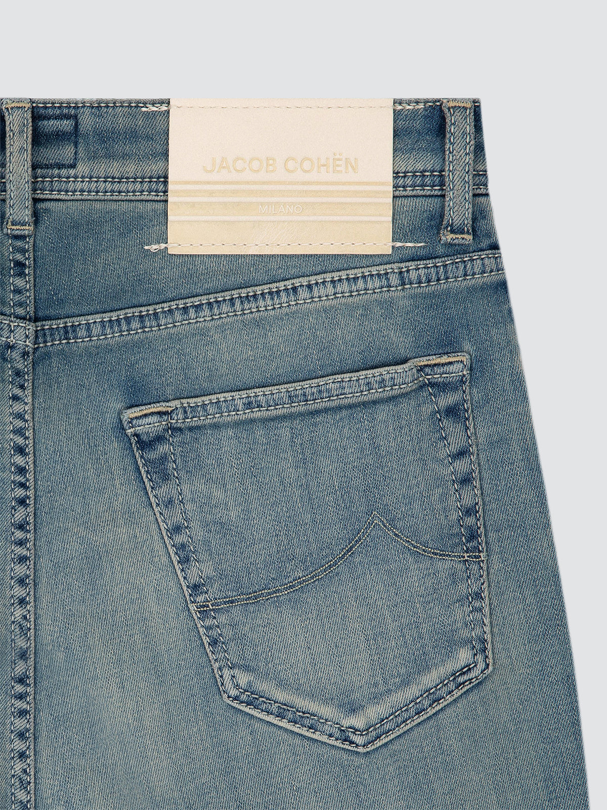 Close-up of the back waistband and pocket of light blue denim jeans, showing a cream-colored fabric brand label stitched onto the back waistband. The brand name 'JACOB COHEN' is printed on the label.