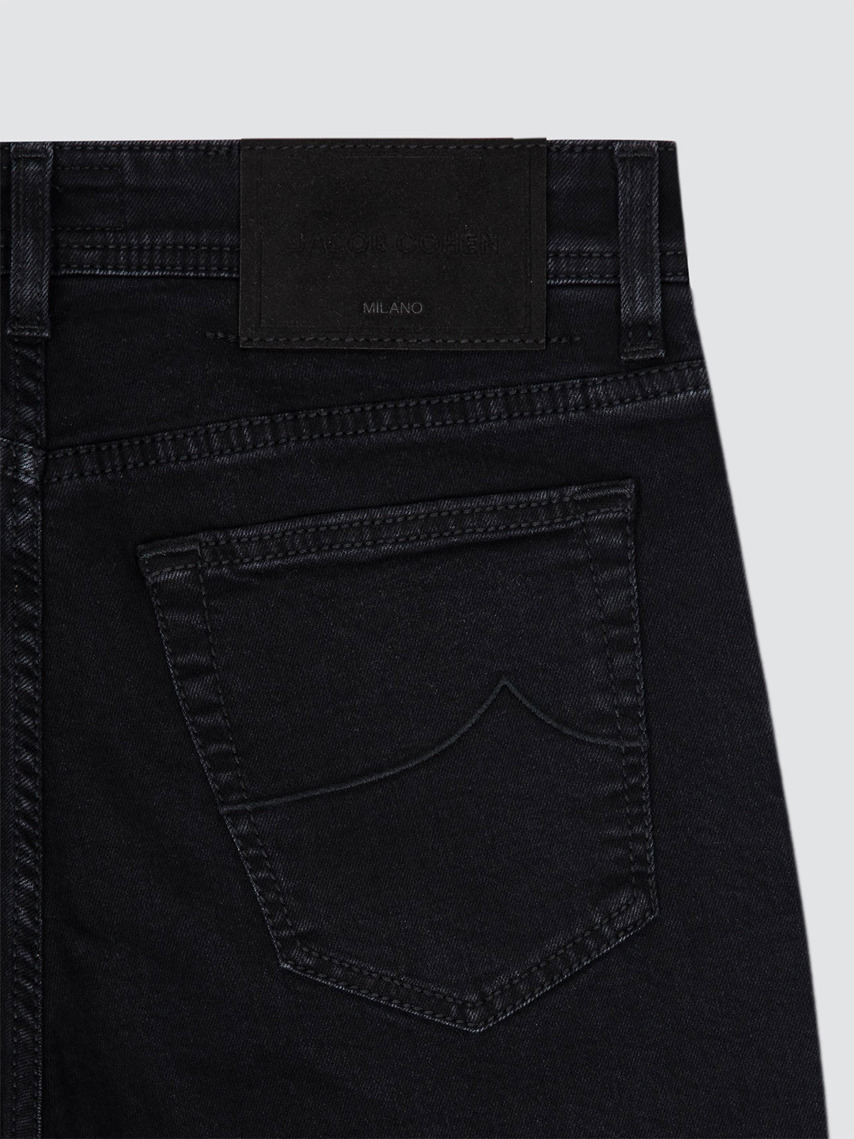 Close-up of the back waistband and pocket area of black denim jeans, showing a black leather patch with 'JACOB COHEN MILANO' branding on a light gray background.