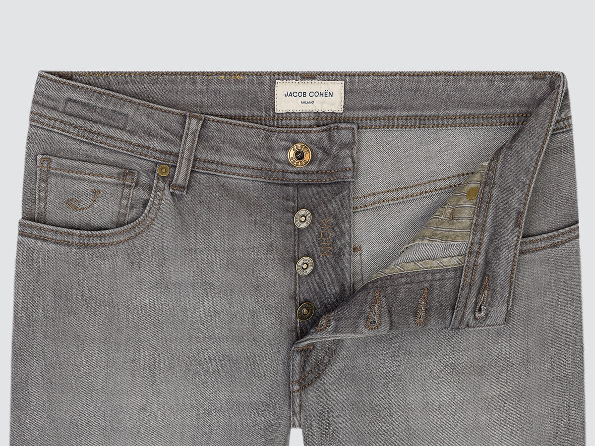 Close-up of gray denim jeans showing the front waistband, button closure, pocket details, and a decorative embroidered patch on the right side. The brand label reads JACOB COHEN.