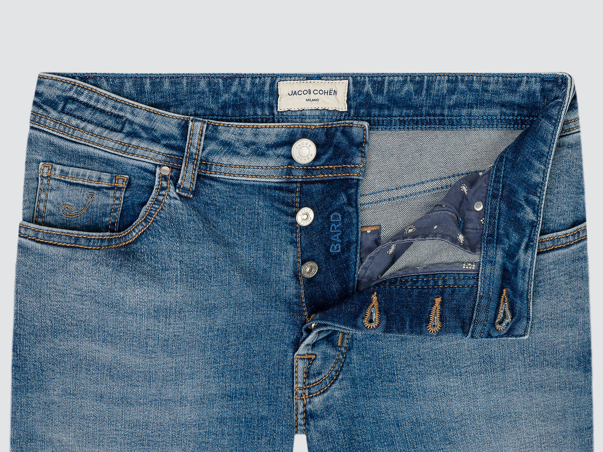 Close-up of blue denim jeans showing the waistband, front button closure, pocket details, and an open back pocket with a patterned lining. A visible waistband label reads Jacob Cohen Milano, and 'Bardo' is printed near the button.