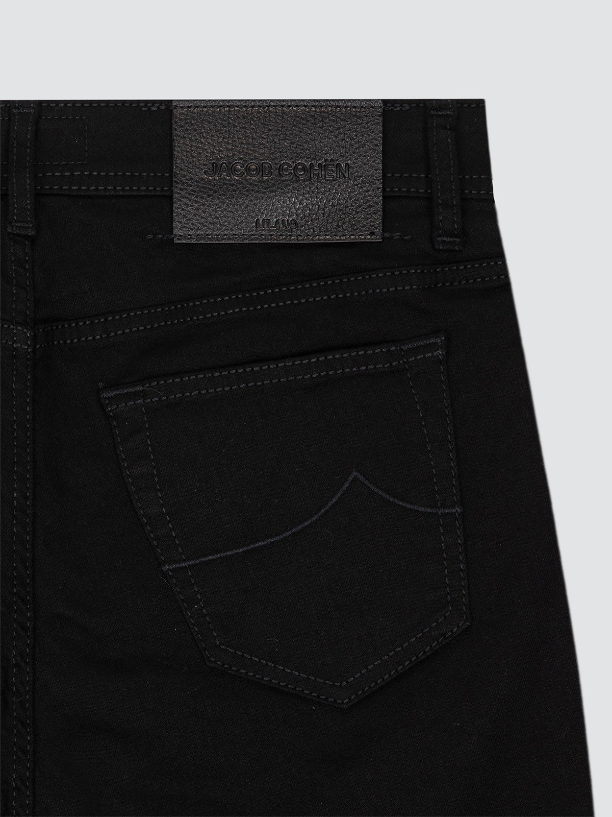 Close-up of the back waistband and pocket of black jeans, showing a dark gray textured brand patch with embossed text reading 'Jacob Cohën'.