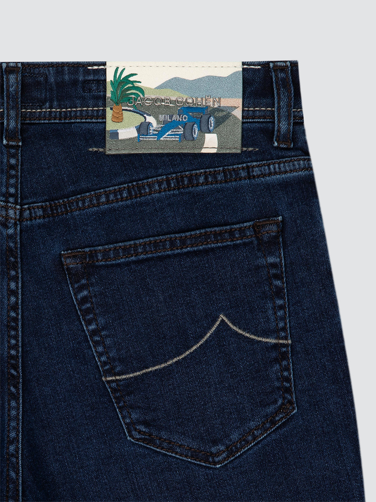 Close-up of the back waistband of dark blue denim jeans, showing a colorful embroidered brand patch formula 1 with a blue car and the text 'Jacob Cohën' and 'MILANO'.