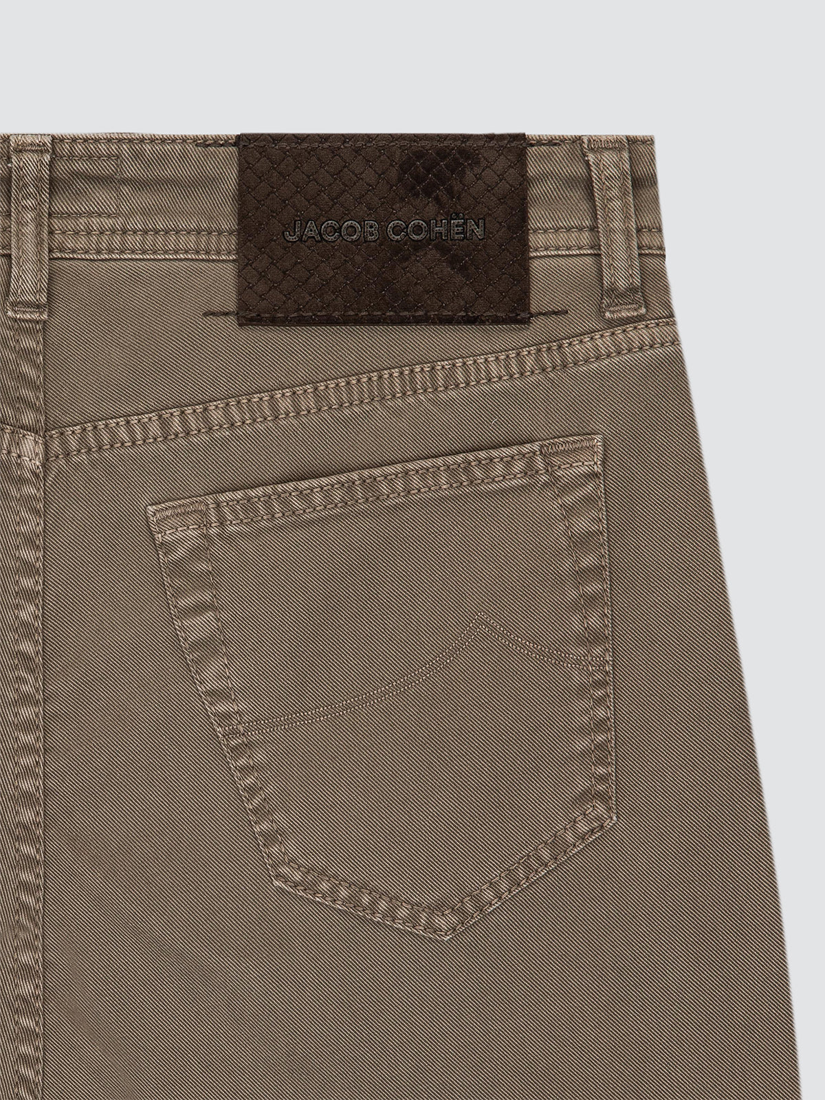 Close-up of the back waistband and pocket of a pair of brown denim jeans, showing a dark brown woven brand patch with 'JACOB COHEN' on a light gray background.