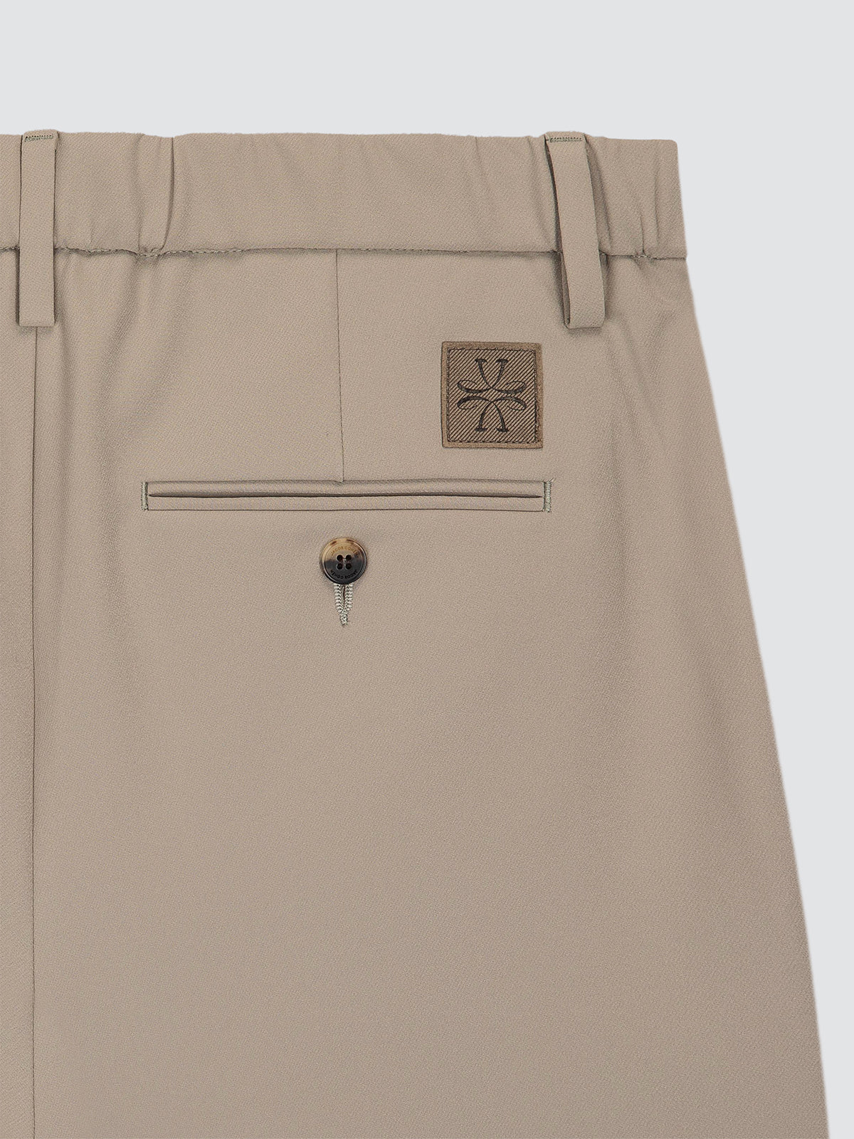 Close-up of the back waistband and rear pocket of beige trousers, showing belt loops, a welt pocket, a decorative button, and a small square patch with a stylized cross logo.