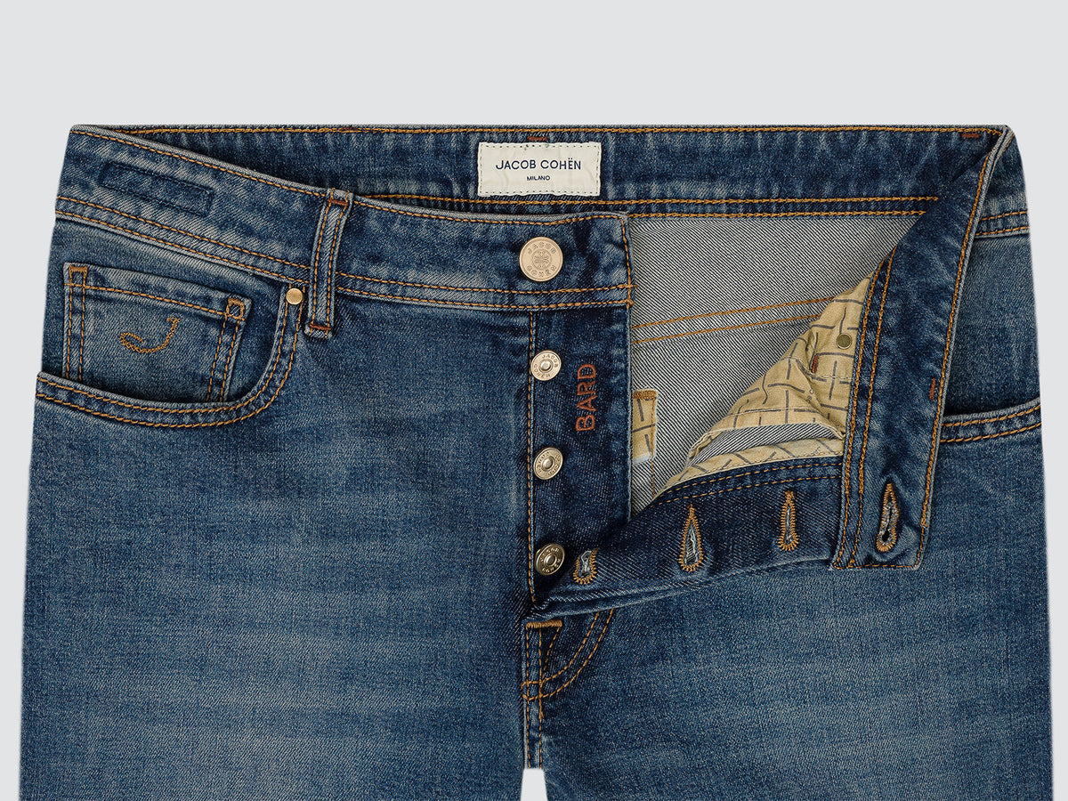 Close-up of the open button fly of medium blue Bard jeans, showing the flat-top branded button and the patterned inner facing.