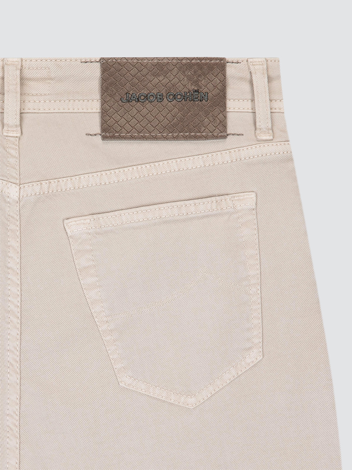Close-up of the back waistband of light beige cotton 5 pockets, showing a brown woven brand patch with 'JACOB COHEN' embroidered on it, and a single back pocket.