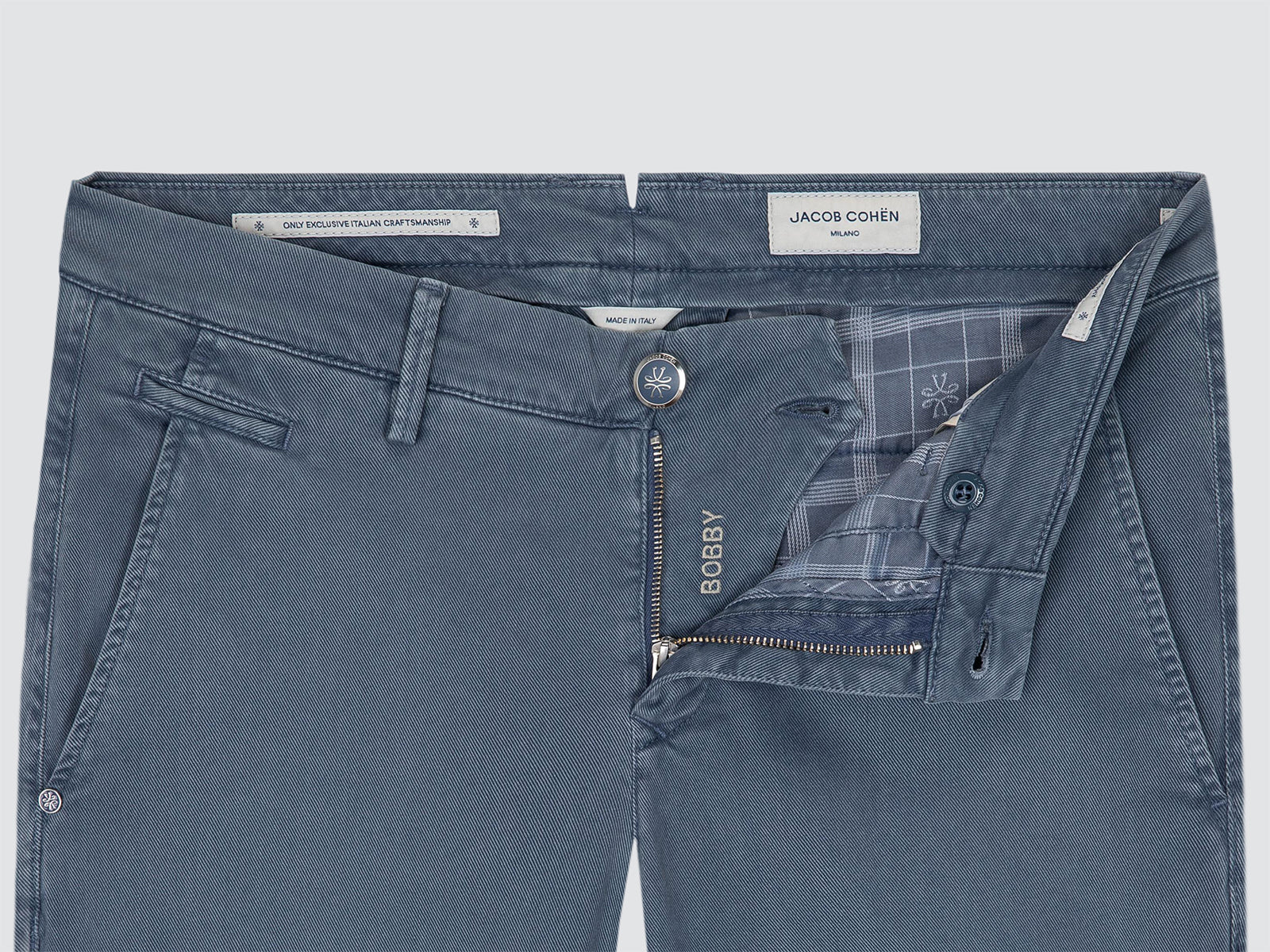 A close-up of a pair of blue chino by Jacob Cohen, showing the front waistband, button closure, zippered fly, and interior plaid lining. The brand name 'Jacob Cohen' is visible on the inner label, and 'BOBBY' is embroidered on the fly zipper pull.
