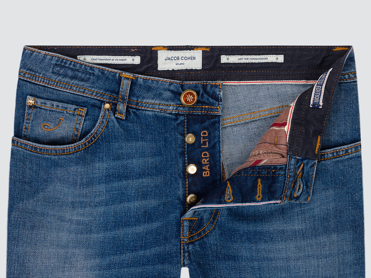 Close-up of the open button fly of mid blue selvedge denim jeans, showing the copper-finish tack button, the striped inner facing and the Bard LTD interior tab.