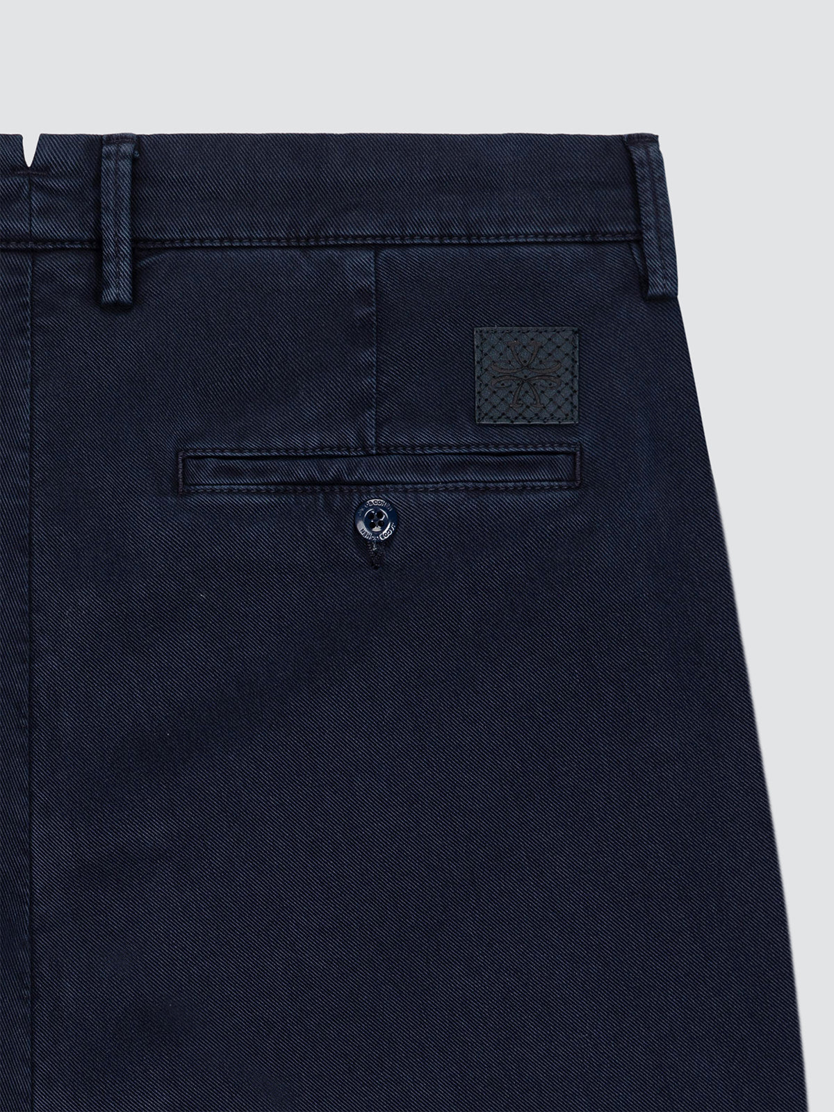 Close-up of the back waistband and pocket area of dark navy trousers, showing belt loops, a buttoned welt pocket, and a small woven brand patch on a light gray background.