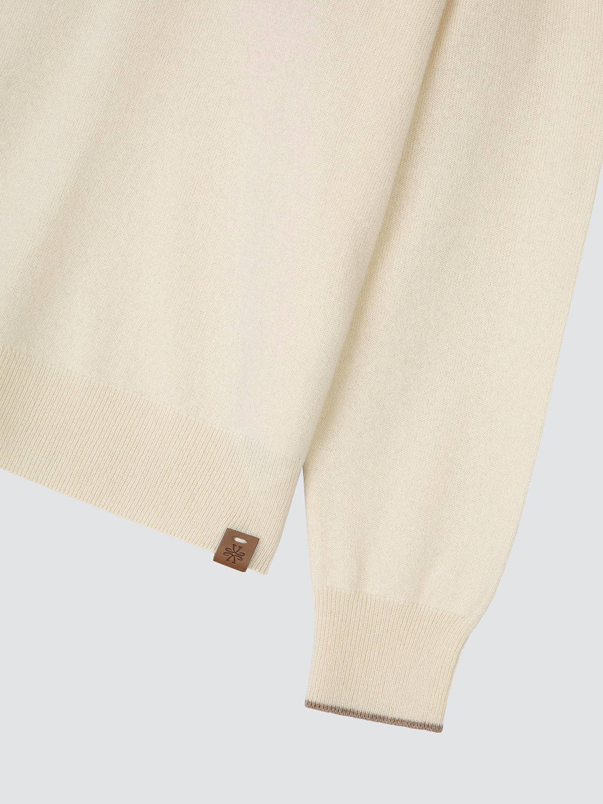 Close-up of the sleeve and ribbed cuff of a cream cashmere Jacob Cohën polo, with a small brown leather brand tab and a subtle contrasting edge along the cuff.