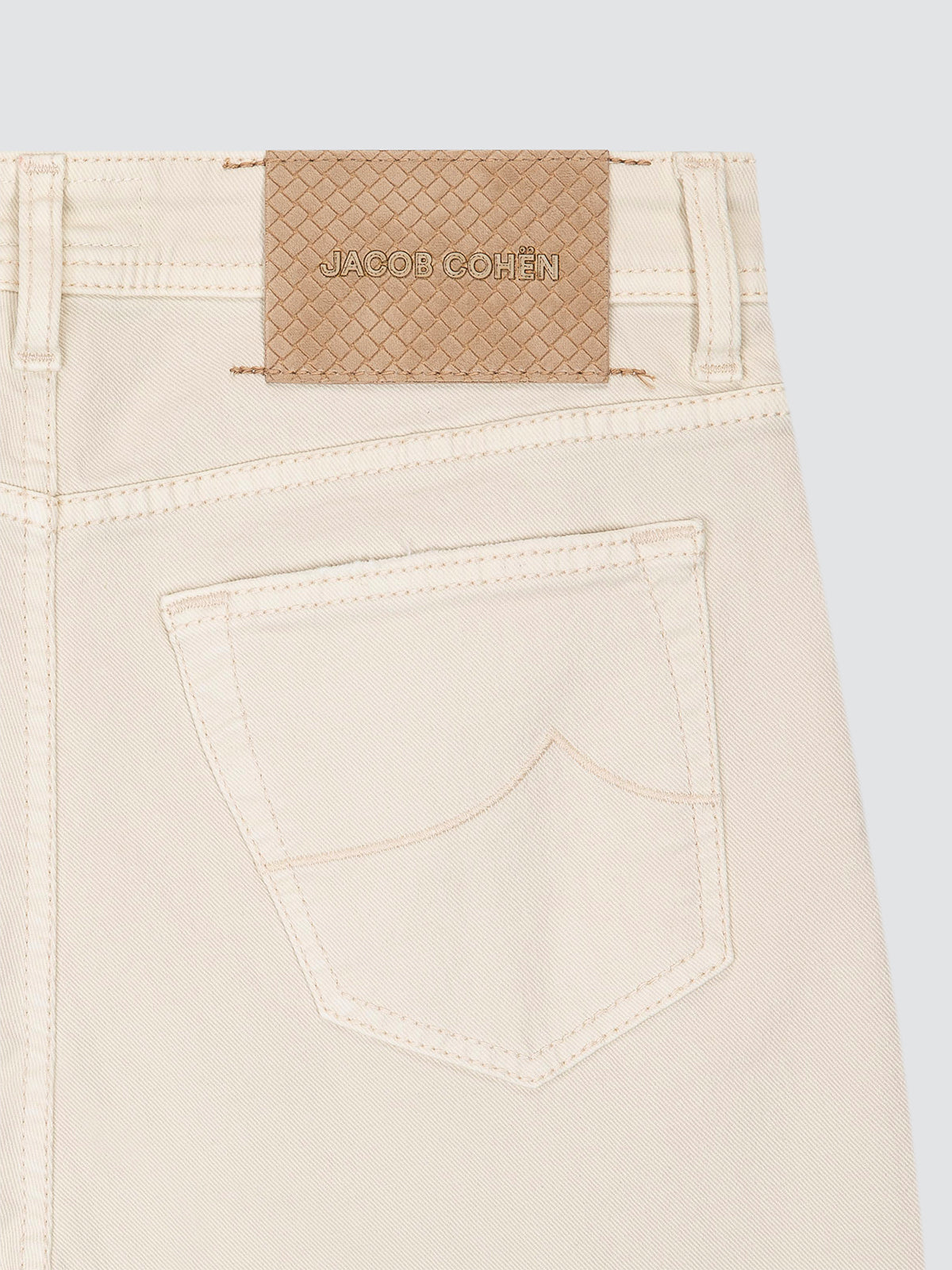 Close-up of the back waistband of light beige denim jeans, showing a woven jacquard brand patch that reads 'JACOB COHEN' on a light gray background.