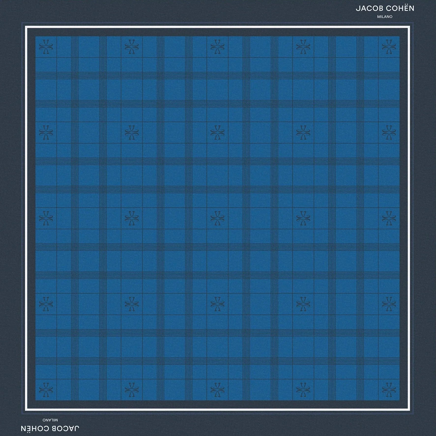 A square scarf featuring a blue and navy tartan plaid pattern, framed by a dark navy border with white text reading 'JACOB COHEN NEW YORK' at the top and bottom, set against a solid beige background.
