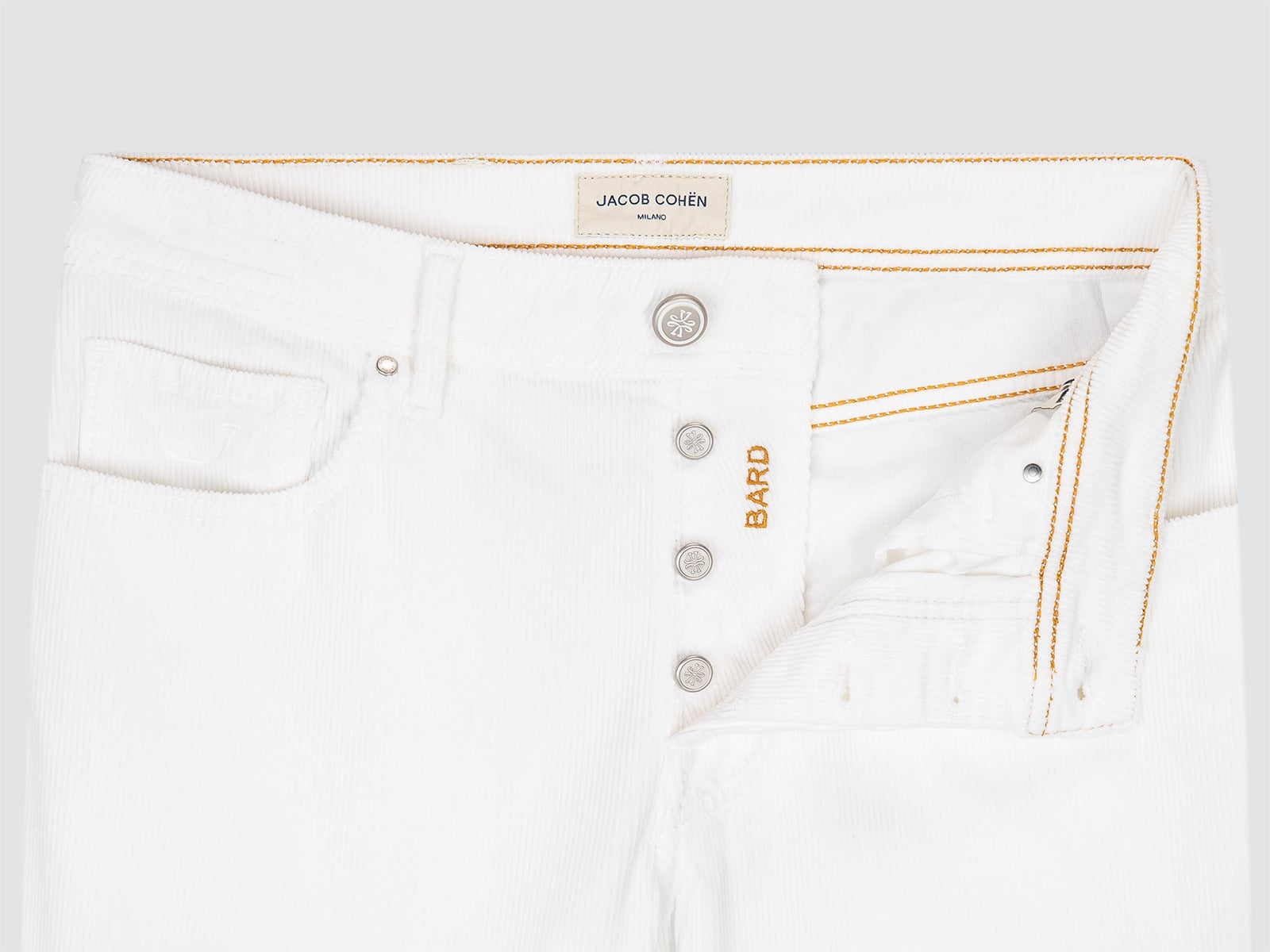 Close-up of white denim jeans showing the waistband, button fly, pocket, and orange contrast stitching. The inner label reads JACOB COHEN, and the side embroidery reads BARD.