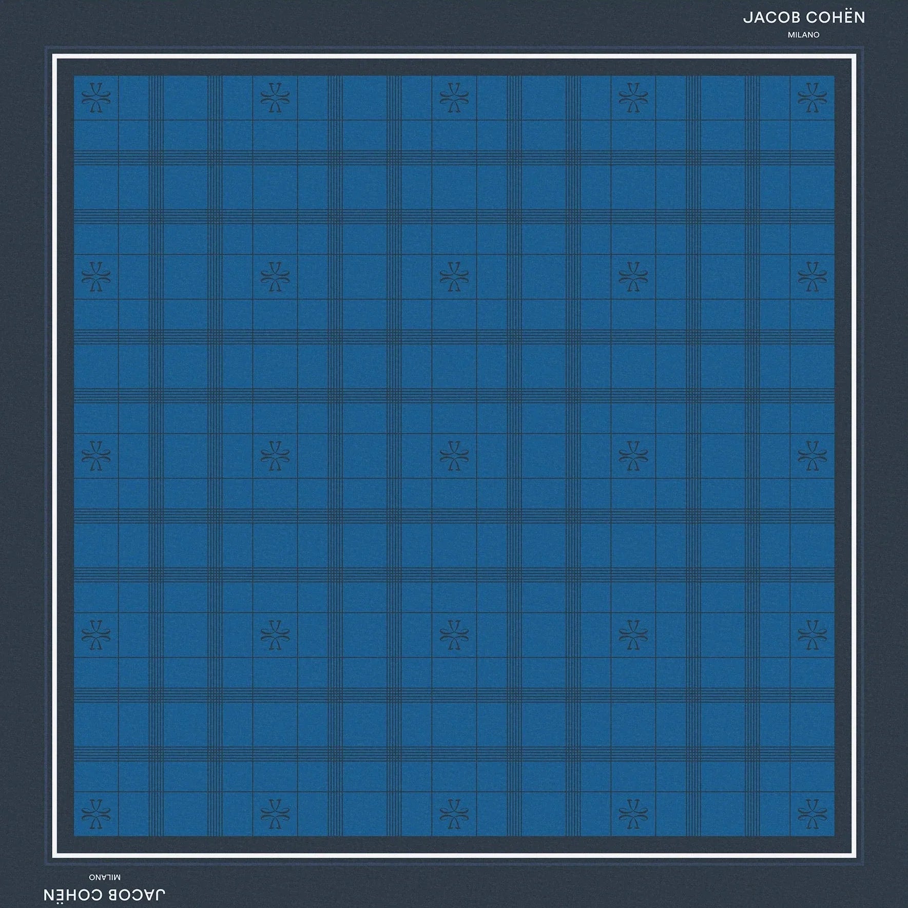 A square scarf featuring a blue and navy tartan plaid pattern, framed by a dark navy border with white text reading 'JACOB COHEN NEW YORK' at the top and bottom, set against a solid beige background.