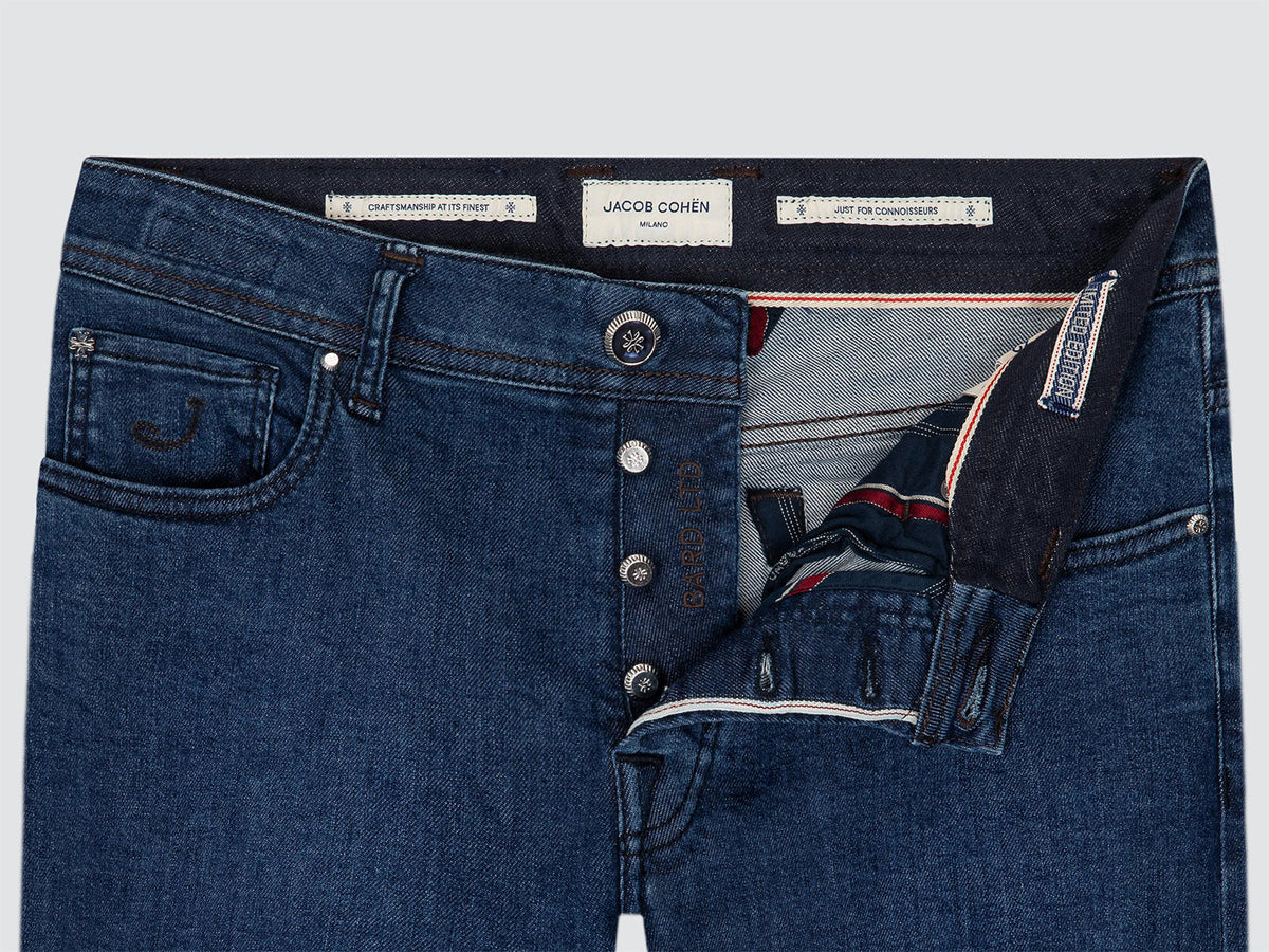 Close-up of the open button fly of the Bard LTD jeans by Jacob Cohën, showing the striped inner lining, the branded waistband labels and the tack button.