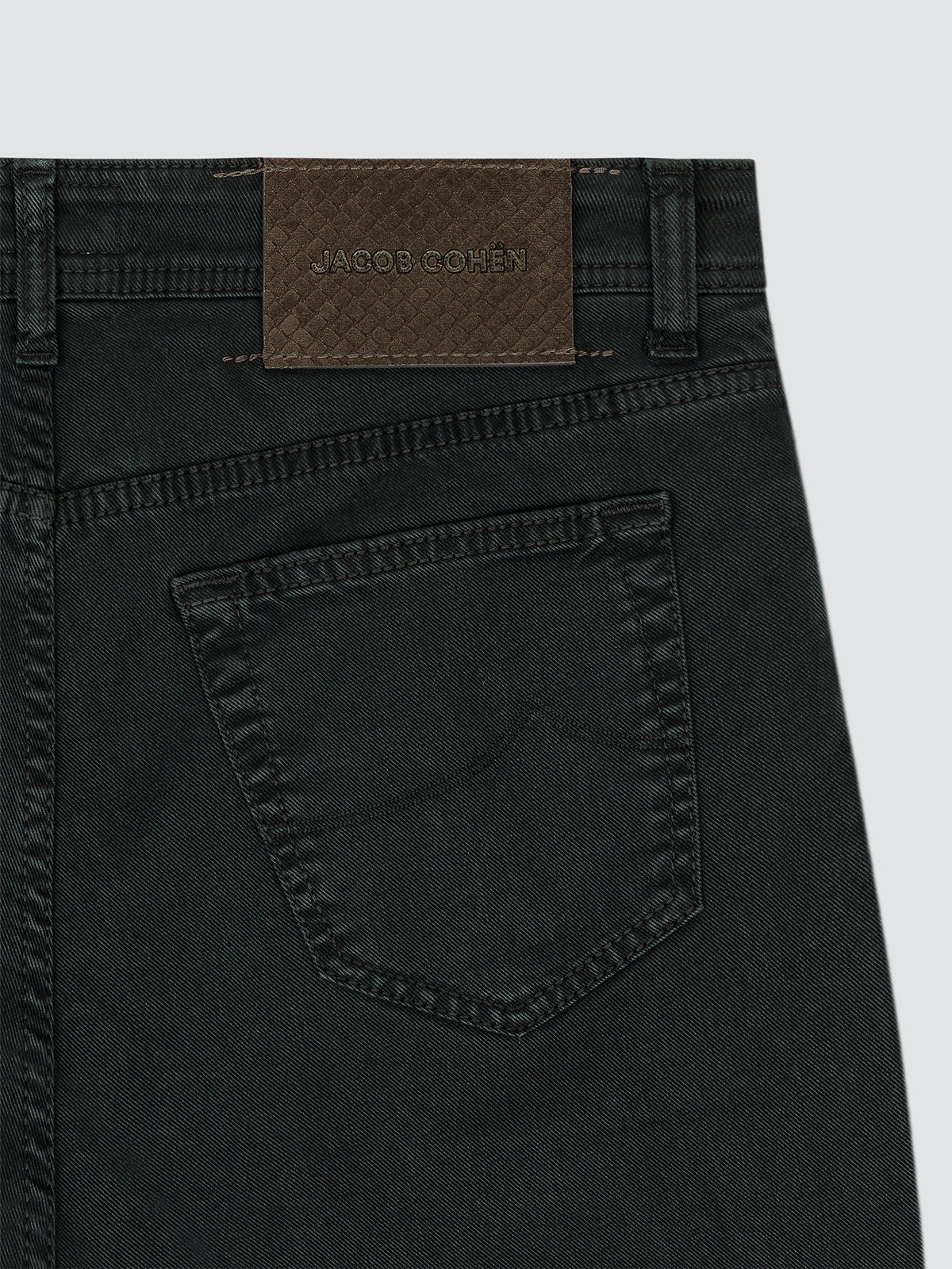 Close-up of the back waistband and pocket of dark washed denim jeans, showing a brown woven brand patch with 'JACOB COHEN' on a light gray background.
