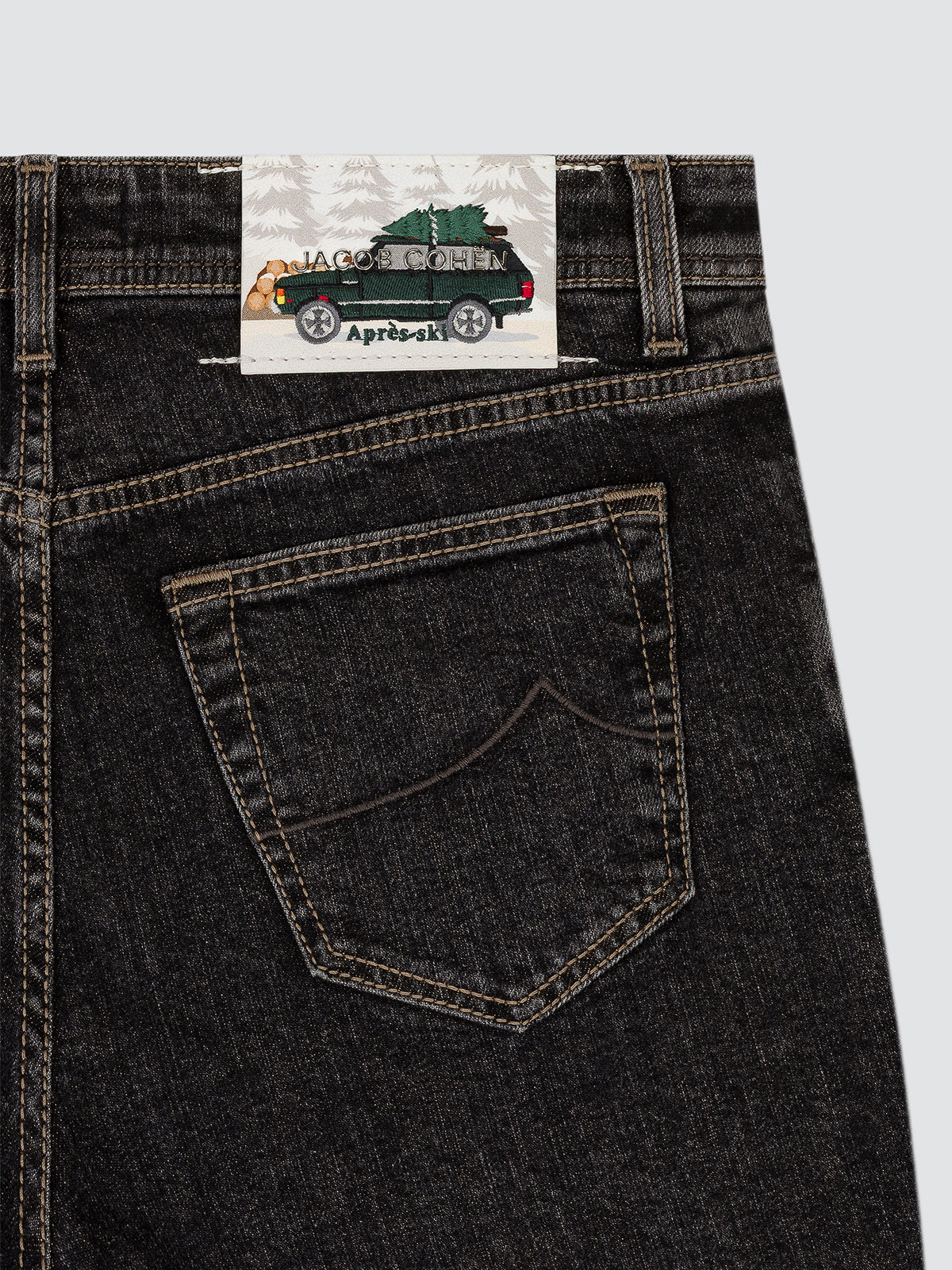 Close-up of the back waistband and pocket of dark washed denim jeans, showing a paper brand label with a green vehicle illustration and the text 'JACOB COHEN.