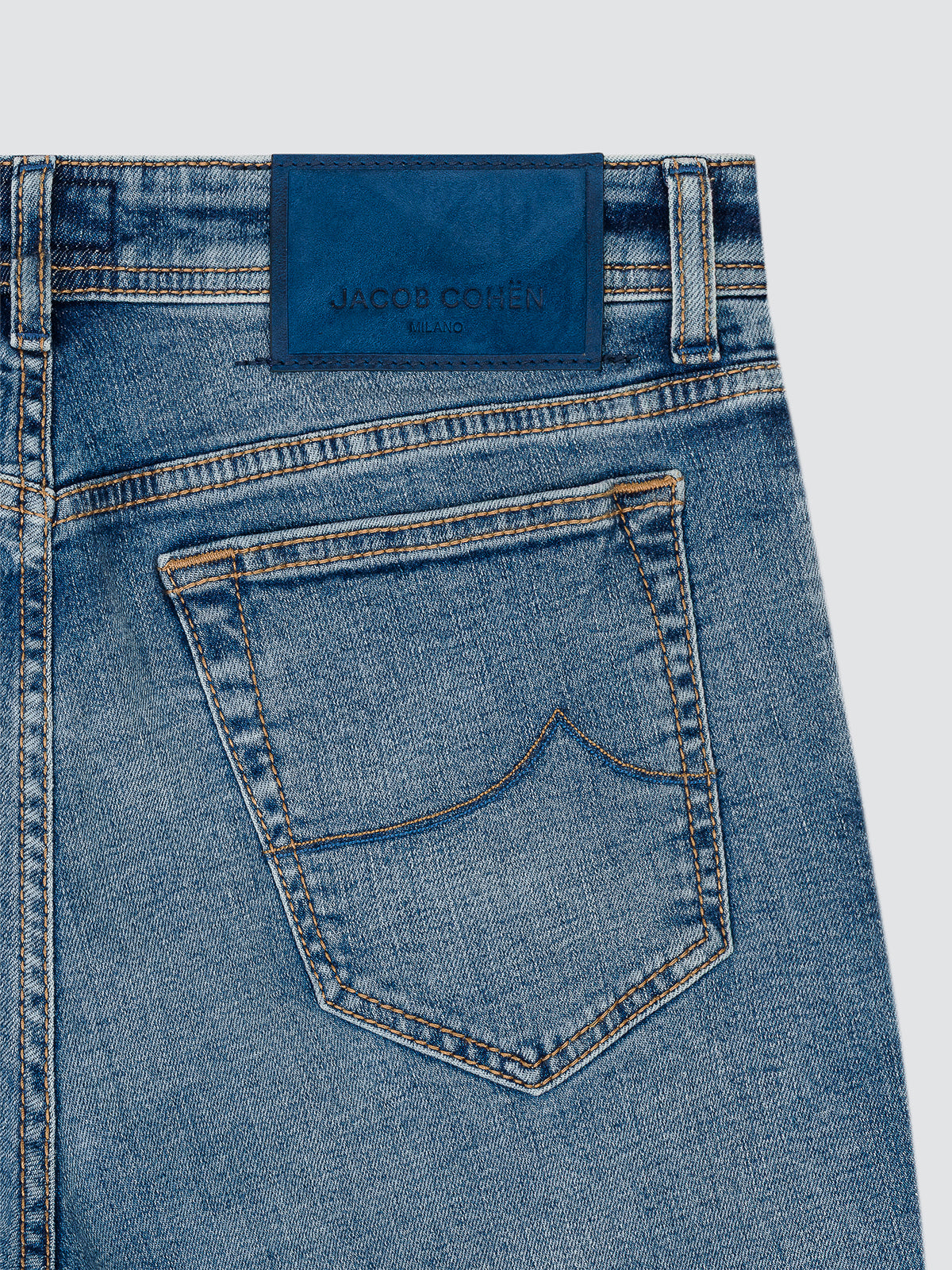 Close-up of the back waistband and pocket of blue denim jeans, showing a dark blue leather patch with the embossed brand name 'JACOB COHEN'.