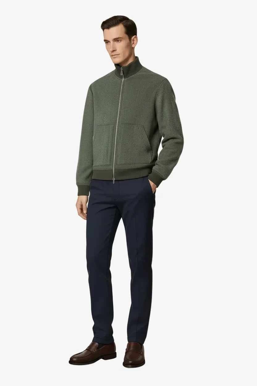 A man modeling a dark olive green zip-up jacket with a high collar, paired with navy blue trousers and brown leather shoes, against a solid light beige background.