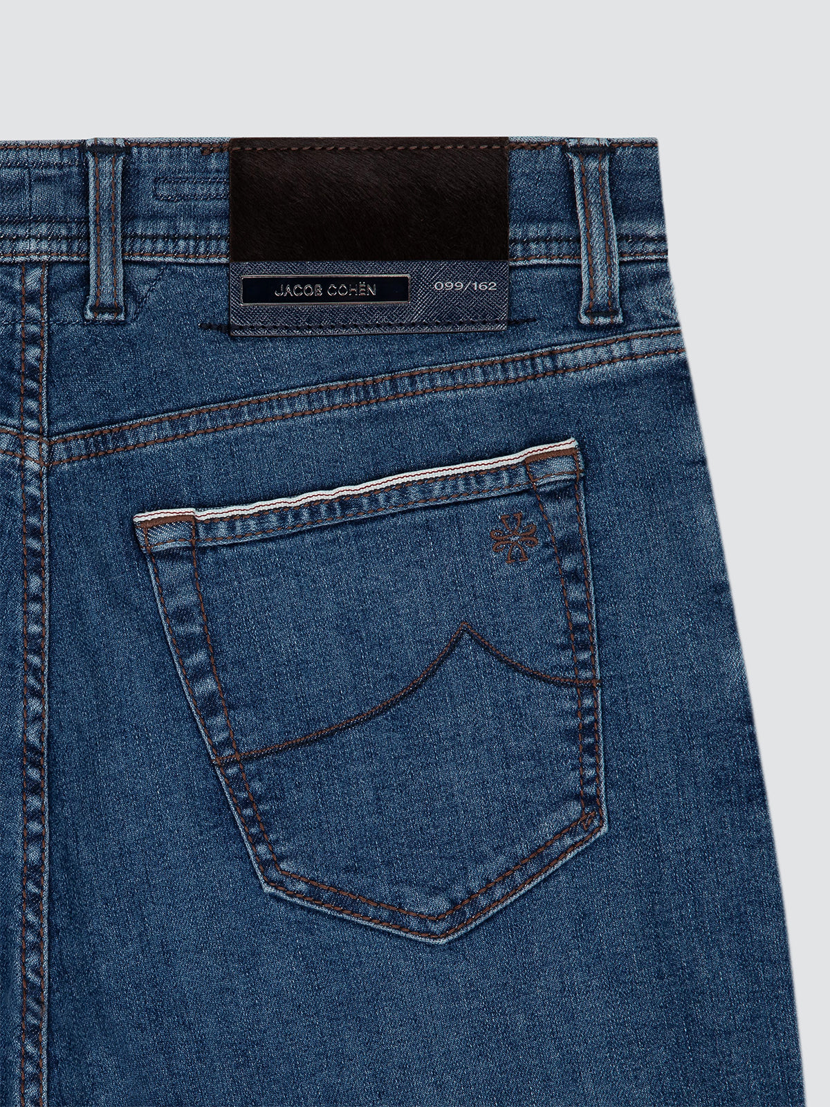 Close-up of the back waistband and pocket area of blue denim jeans, showing a dark brown leather brand patch that reads ltd JACOB COHEN and a small embroidered logo on the pocket.