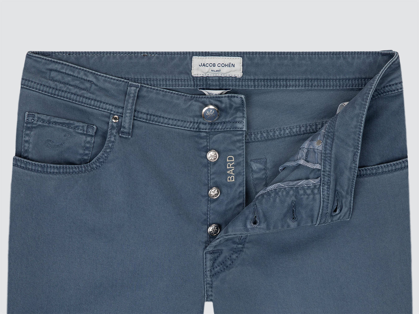 Close-up of a pair of blue denim jeans showing the waistband, front button closure, and pocket details. The brand label 'JACOB COHEN' is visible at the back waistband, and 'BARD' is embroidered on the front button placket.