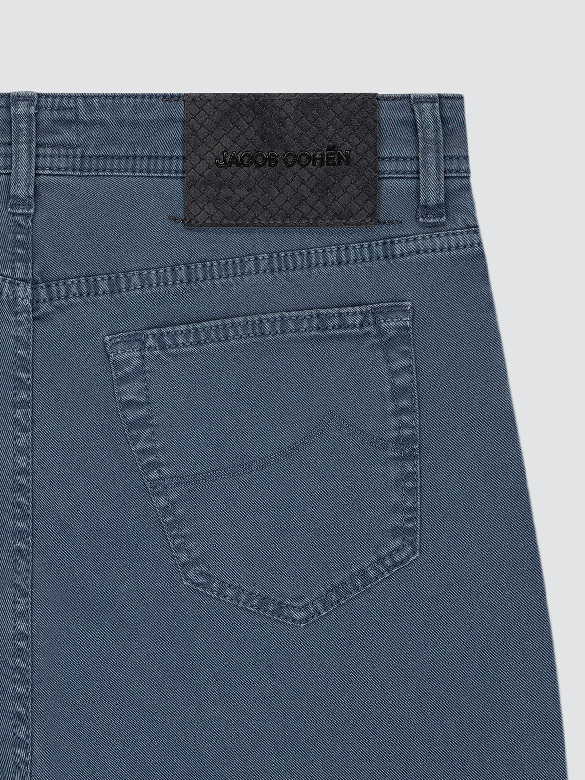Close-up of the back waistband and pocket of blue denim jeans, showing a dark woven brand patch with the text JACOB COHEN.