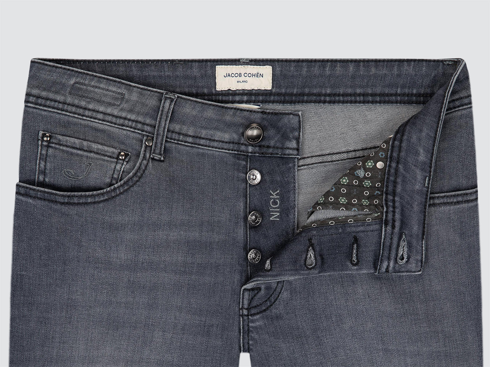 Close-up of dark gray denim jeans showing the front waistband, button closure, pocket, and decorative studded detail on the right side. The inner label reads NICK, and the waistband label shows JACOB COHEN.