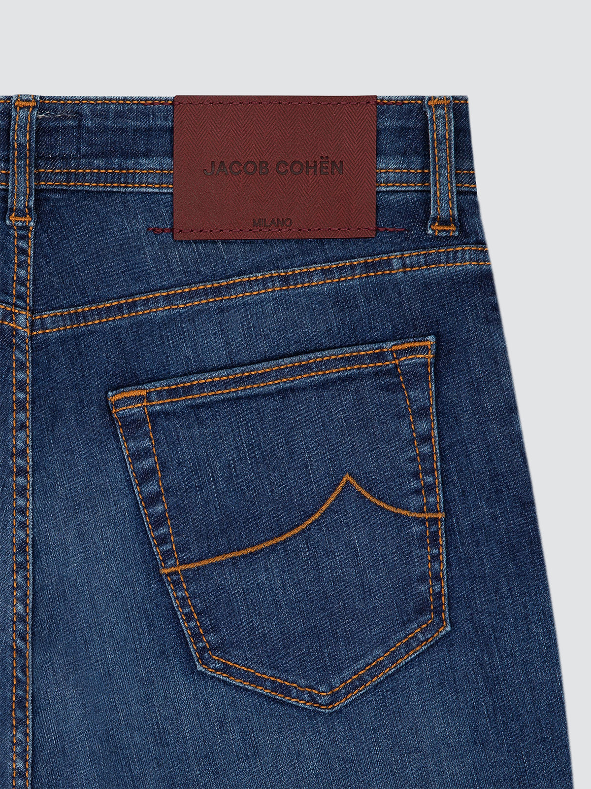 Close-up of the back waistband and pocket of blue denim jeans with orange contrast stitching and a brown leather brand patch reading JACOB COHËN.