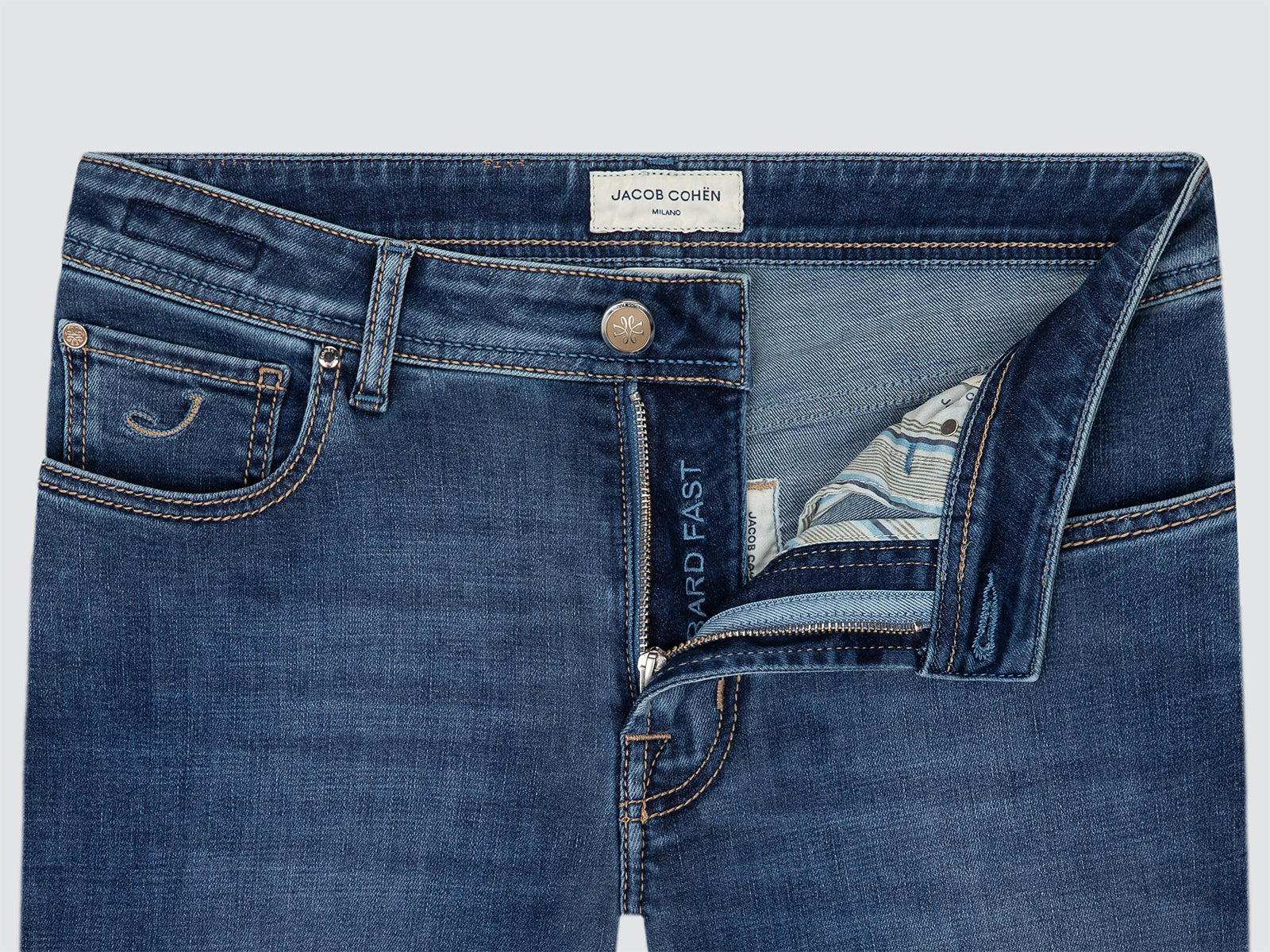 Close-up of a pair of blue denim jeans showing the waistband, front pocket, button, and partially unzipped fly with a patterned lining. The inner label reads JACOB COHEN and the side text reads BARD FAST.