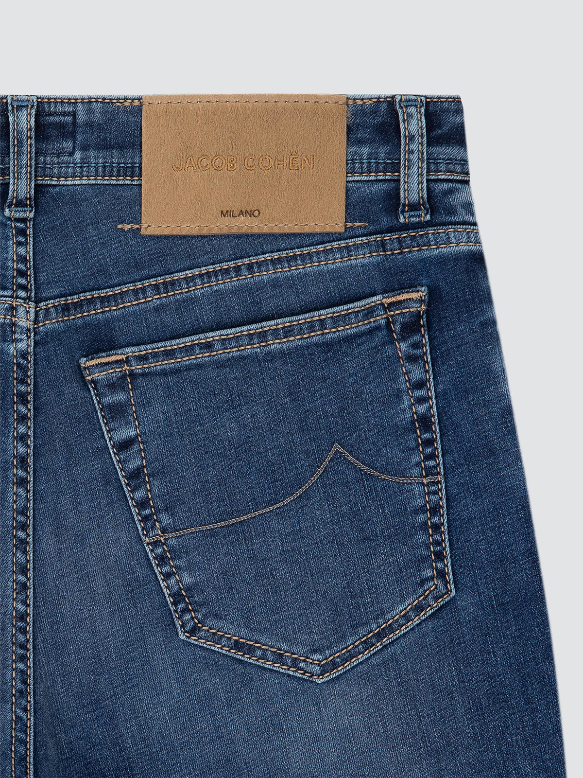 Close-up of the back waistband and pocket area of blue denim jeans, showing a tan leather patch with the brand name Jacob Cohën and 'Milano' on a light gray background.