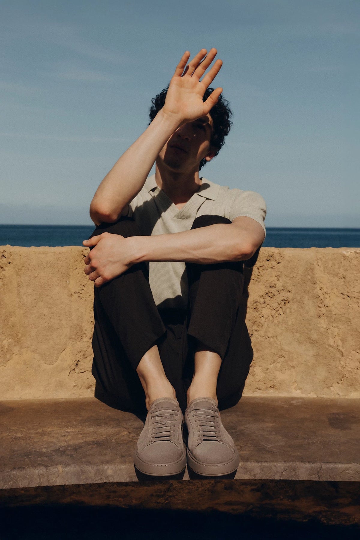 Hombre sentado en un muro frente al mar, con polo blanco, pantalón negro y zapatillas Santoni grises